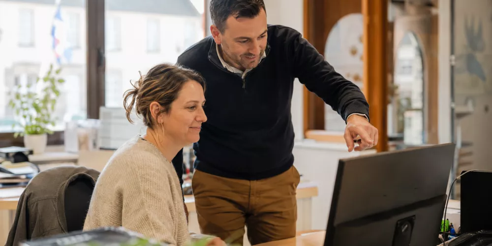Deux agents d’une collectivité locale travaillent ensemble devant un ordinateur dans un bureau municipal lumineux, avec une ambiance chaleureuse et professionnelle représentative des mairies françaises